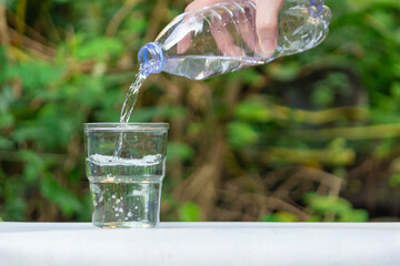 A hand pours clear, pure drinking water from a plastic bottle into a transparent glass, set against a blurred natural green background. The scene evokes freshness, hydration, and healthy living.