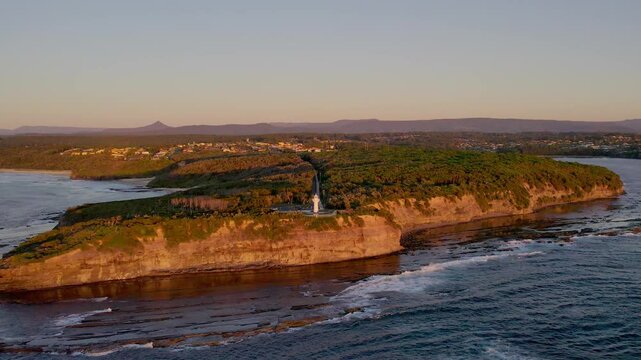 Calm view of the rocky coastline near Ulladulla, with Warden Head Lighthouse bathed in warm morning light. The peaceful scene highlights the rugged beauty and vastness of the coast.