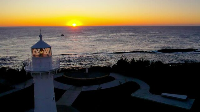 Drone flies toward the horizon over Warden Head Lighthouse at sunrise near Ulladulla, NSW. A fishing boat cruises through the calm ocean, adding life to this peaceful coastal morning scene.
