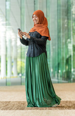 Smiling Asian muslimah using smartphone indoors with green outdoor view. Confident Muslim woman...