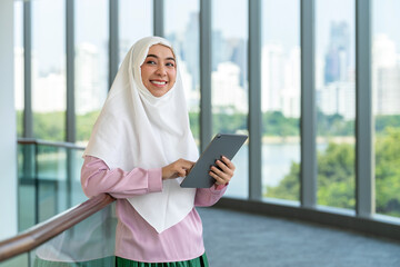 Smiling Asian muslimah using tablet indoors with city skyline background. Confident Muslim woman...