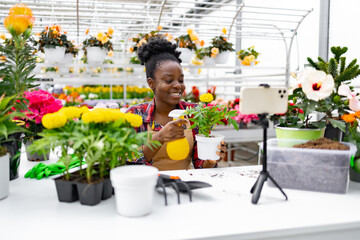 An African American woman smiles while spraying a plant, with a phone recording. Surrounded by various colorful flowers.