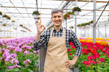 A friendly man in a greenhouse gives an 'okay' sign, surrounded by vibrant flowers, indicating...