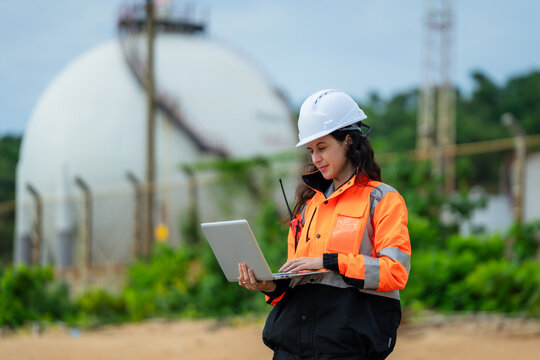 Female engineer concentrating on data analysis on a laptop at a natural gas storage facility. A woman inspector in safety gear working on a computer at an oil and gas refinery plant.
