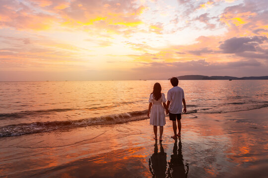 Young happiness Asian couple traveler relaxing and watching beautiful Sunset on the tranquil beach, Traveling on summer vacation concept