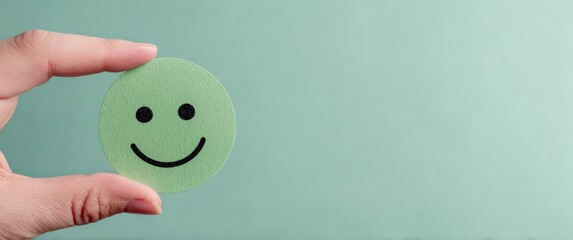 Hand holding a green smiling emoji on a light mint background, conveying a concept of happiness and positive emotion, with copy space