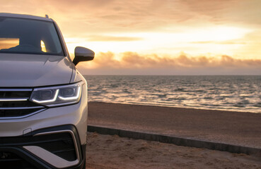 Silhouetted by a colorful sunset, a gray car stands near the ocean's edge. The warm light and open...