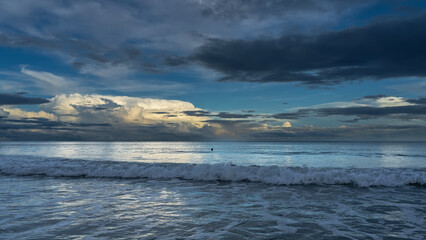 Early morning by the ocean. The waves roll towards the shore, foam and spread. A swimming man's head is visible in the distance. The clouds in the blue sky are illuminated with golden light.Reflection