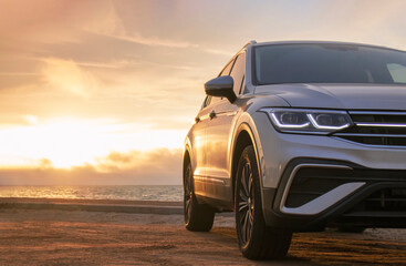 Silhouetted by a colorful sunset, a gray car stands near the ocean's edge. The warm light and open...