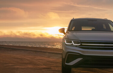 Silhouetted by a colorful sunset, a gray car stands near the ocean's edge. The warm light and open space capture a moment of solitude and exploration. 