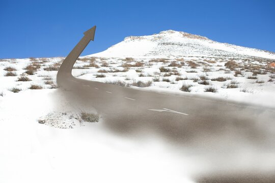 Fototapeta Snowy scene showing paved road emerging into 3D arrow against clear blue sky, dry shrubs