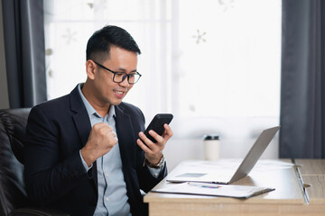 Young businessman celebrating success while using smartphone in modern office environment with laptop on wooden desk and natural light coming through the window © WMSTUDIO