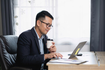 Professional man working on laptop with coffee cup in modern office, focusing on tasks, wearing glasses, sitting at wooden desk with papers and documents