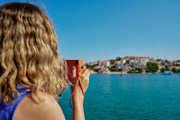 Woman standing near sea holding instant camera and taking photo. Female tourist at coastal resort...