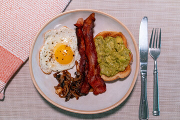 Hearty breakfast plate with crispy bacon, fried egg, sautéed mushrooms, and creamy avocado toast on rustic ceramic dish—perfect for brunch menus, food blogs, or nutrition features.