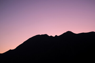 Glowing twilight sky blankets the rolling hills of Sembalun, Lombok, painting the horizon in soft purples and oranges as mountain silhouettes fade into serene dusk.