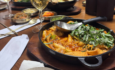 Close-up of Korean tteokbokki served in a hot pan with ramen noodles, fish cakes, and fresh perilla leaves. Surrounded by side dishes and wine on a wooden table in a cozy restaurant setting.