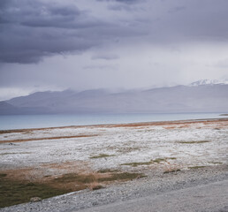 Rocky shore of Lake Karakul with blue water and mountain peaks in the background, cloudy weather...