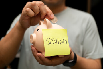 Man Depositing Coin into Piggy Bank with Saving Note. A man's hand is shown actively depositing a...