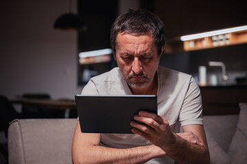 Tired exhausted man reading social media content on digital tablet computer at home late at night