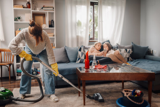 Mother vacuuming living room while daughters relaxing on sofa