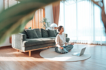 Happy young Asian woman practicing yoga and meditation at home sitting on floor in living room in lotus position with laptop