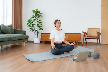 Young Asian woman meditating in lotus position at home, sitting on the floor in fitness clothing. Breathing workout to achieve relaxation, peace and mindfulness
