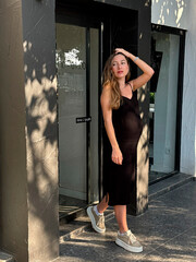 Woman standing at modern building entrance wearing black dress and casual shoes in sunny weather