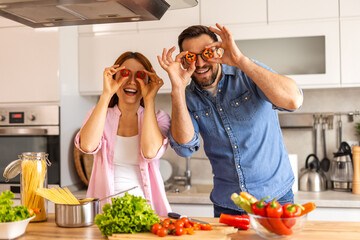 Cheerful couple enjoys a playful moment in the kitchen holding sliced vegetables over their eyes. kitchen is filled with fresh ingredients like lettuce, tomatoes, peppers, and pasta