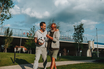 Sporty father and adult son walking home after football match in park.