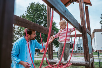 Toddler crossing playground obstacle with dad's support