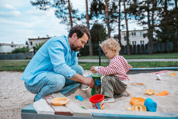 Father and little toddler son playing in the sandbox on playground.