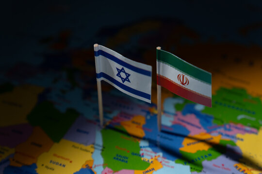Miniature flags of Israel and Iran are planted on a blurred map. International relations, conflict, diplomacy, and geographical focus on the Middle East concept.
