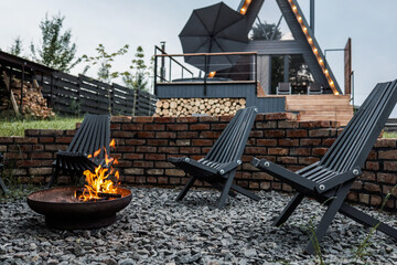 Burning firewood in a metal firepit surrounded by gravel and black wooden chairs, with a modern...