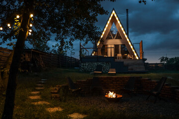 Illuminated A-frame cabin glowing in the dark with a cozy campfire and garden lights, surrounded by trees and dramatic evening sky