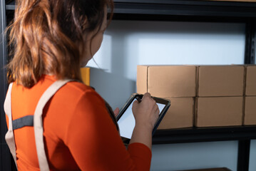 A woman uses a digital tablet to check stock inventory at home, She stands next to shelves with cardboard boxes, representing a small business operation.