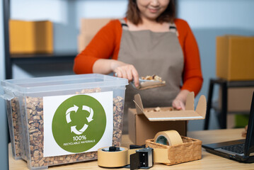 An Asian woman is scooping eco-friendly protective packaging made from water hyacinth into a cardboard box — a sustainable packaging practice that helps reduce plastic waste.