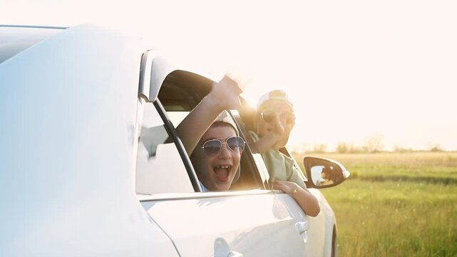 Two little cheerful boys drive away in a car along a country road at sunset, shouting, smiling and waving hands goodbye. Family trip and vacations on the nature in summer, happy childhood