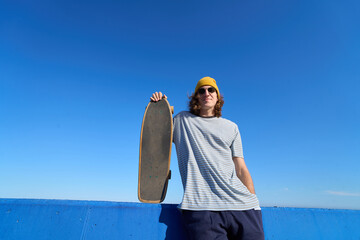 Skater holding skateboard against blue wall and sky