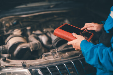 A mechanic uses a diagnostic tablet to inspect a car engine under the hood in a workshop setting.