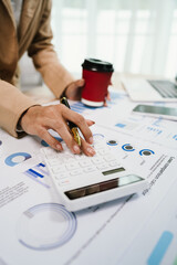 Close-up of businesswoman analyzing chart on clipboard with pen, surrounded by technology such as laptop and smartphone, in busy work environment.