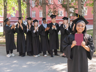 Young African woman holds diploma while classmates applaud her. 