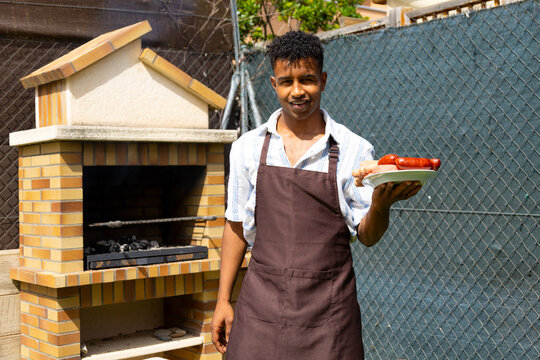 Young man holding grilled sausages on plate in garden with barbecue