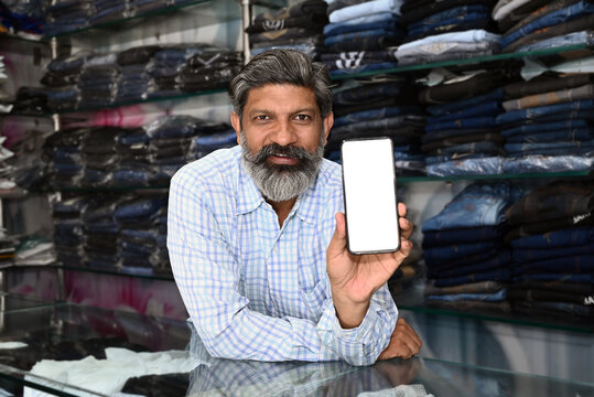 Indian smiling merchant in clothing store hold mobile with white screen mock up by looking at camera - concept of Technology, advertisement, online booking and e-commerce and product promotions 