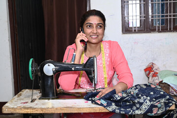 Indian rural woman talking on phone while stitching clothes with sewing machine at home 
