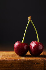 Two juicy cherries or sweet cherries on one branch covered with water droplets. Dark background, wooden stand. Space for text. Summer berries and fruits