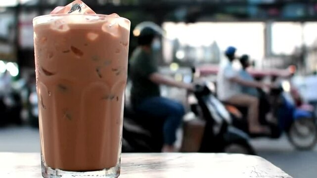 An iced drink in a glass possibly Thai iced tea sits on a table with blurry street scene of vehicles  buildings (AI Video)