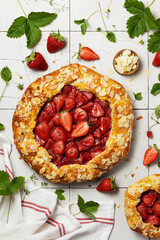Shortcrust pastry galette with strawberry filling on cracked white tile background. Round fruit pie with almond petals sprinkle. Fresh berries and strawberry leaves lie next to the pie. Top view