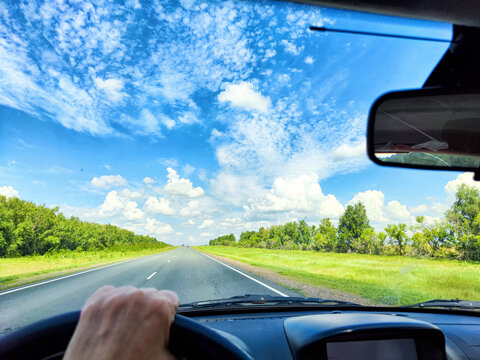 view through car windshield of natural landscape with green field, white clouds in blue sky. Hand of woman on steering wheel of car. Female traveler