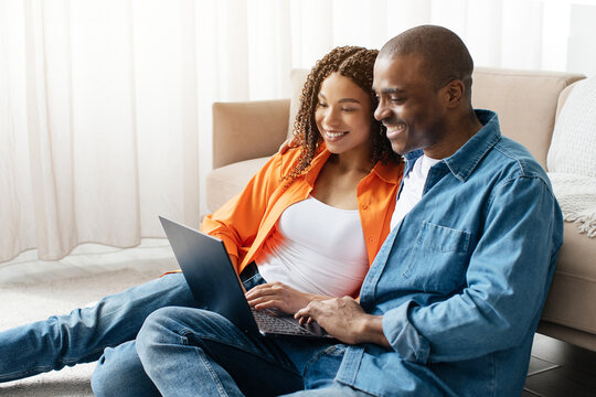 Happy couple sitting on floor, browsing laptop together and smiling, cheerful african american spouses enjoying relaxed weekend moment at home in modern interior, closeup - Powered by Adobe